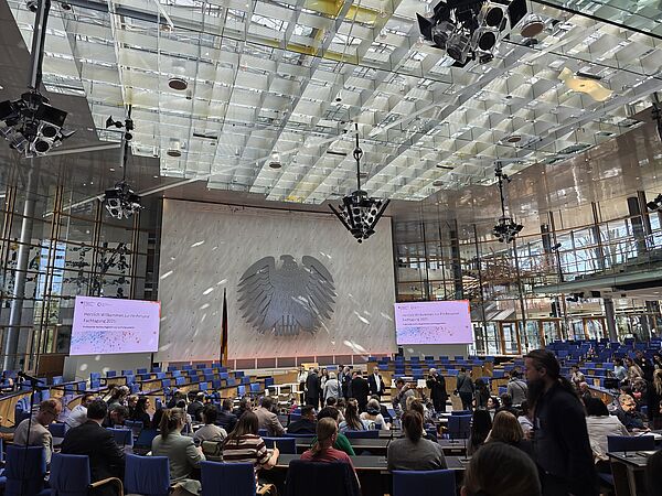  Großer Konferenzsaal mit Bundesadler an der Wand, zwei Leinwänden mit Begrüßungstext und vielen sitzenden sowie stehenden Menschen.