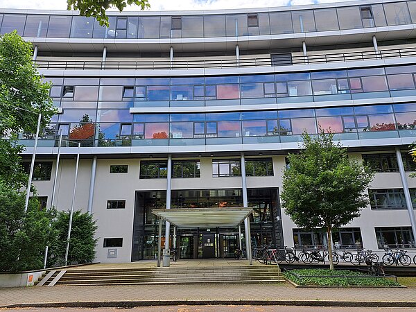 Ein siebenstöckiges, weißes Gebäude mit vielen großen Fenstern und zum Teil auch einer Glasfassade. Eine kleine, breite Treppe führt zum Haupteingang des Gebäudes. Vor dem Gebäude stehen einige Bäume und viele Fahrräder.