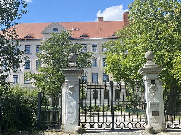 Robert-Schumann-Gymnasium Ein alt verziertes, weißes Gebäude mit rotem Dach. Vor dem Gebäude sind einige grüne Bäume und ein Metallzaun mit zwei weißen Steinpfeilern zu beiden Seiten des integrierten Metalltors.