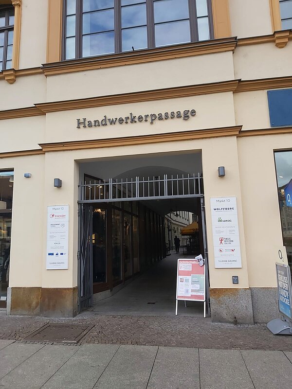 Das Bild zeigt den Eingang zur "Handwerkerpassage" in einem historischen Gebäude. Über dem Torbogen ist der Name "Handwerkerpassage" in großen, metallisch glänzenden Buchstaben angebracht. Es handelt sich um einen Durchgang, der von außen zugänglich ist, und hinter dem Eingang scheint eine schmale Passage mit Geschäften oder Büros zu verlaufen. Links vom Eingang befindet sich ein Schild mit mehreren Logos und Namen, die vermutlich auf Unternehmen oder Institutionen hinweisen, die in diesem Gebäude ansässig sind.
