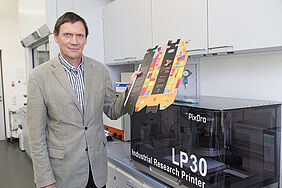 Prof. Eugen Herzau steht neben einer für Forschungszwecke optimierten Digitaldruckmaschine. In der Hand hält er einen digital bedruckten Weinkarton.