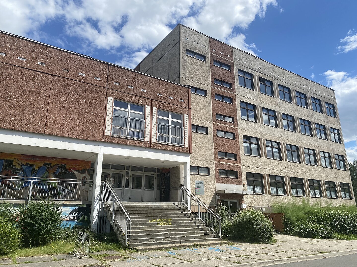 Schule Paunsdorf Das Bild zeigt ein mehrstöckiges Gebäude mit einem modernen, funktionalen Design. Die Fassade ist eine Kombination aus braunen und grauen Wänden mit zahlreichen Fenstern. Der Eingangsbereich hat eine Treppe, die zu doppeltüren führt, und auf beiden Seiten gibt es Geländer. Auf der Wand unterhalb des Eingangs ist Graffiti zu sehen. Das umliegende Gelände ist teilweise bewachsen, und es gibt einen gepflasterten Gehweg.