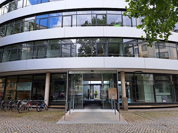 Ein abgerundetes Gebäude mit vielen großen Fenstern. Auf einem Schild über dem Haupteingang steht „Max-Planck-Institut für Kognitions- und Neurowissenschaften“. Auf einem weiteren Schild im Fenster steht „Wissenschaft lebt von Weltoffenheit, Vielfalt und Toleranz!“.