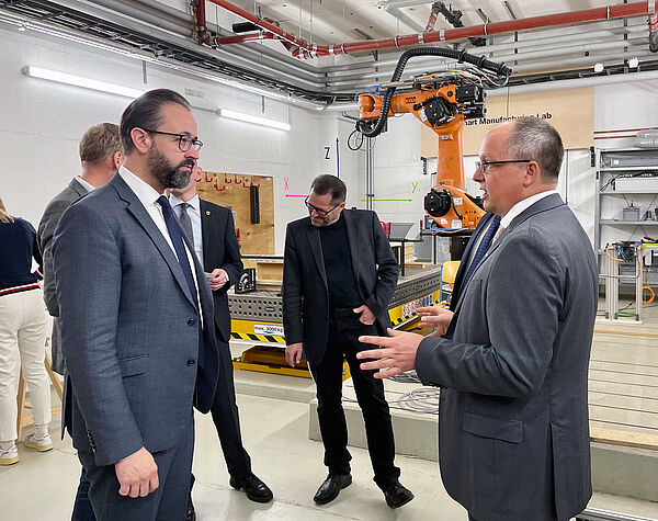 Wissenschaftsminister Sebastian Gemkow und HTWK-Rektor Prof. Mark Mietzner im Gespräch im Smart Manufacturing Lab, im Hintergrund ist ein Roboter zu sehen