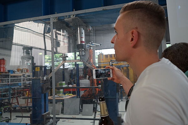 Germo nimmt mit dem Handy in der einen und Bierflasche in der anderen Hand ein Video seiner Brücke im Versuchsapparat auf.