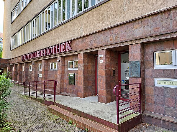 Der untere Teil eines Gebäudes aus roten Steinen und Beton. Eine Rampe mit rotem Geländer führt zum Eingang. Darüber steht in großen roten Buchstaben „Stadtteilbibliothek“.