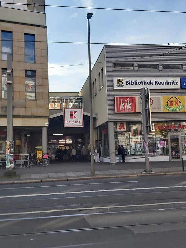 Ein Einkaufszentrum mit verschiedenen Geschäften. An der Fassade hängen unterschiedliche Schilder, darunter auch das der Bibliothek Reudnitz. Vor dem Gebäude befindet sich eine Straße.