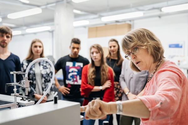 Eine Professorin erklärt Studierenden eine Druckmaschine. Dazu steht dort geschrieben: "Gestalten Sie die Zukunft mit!"