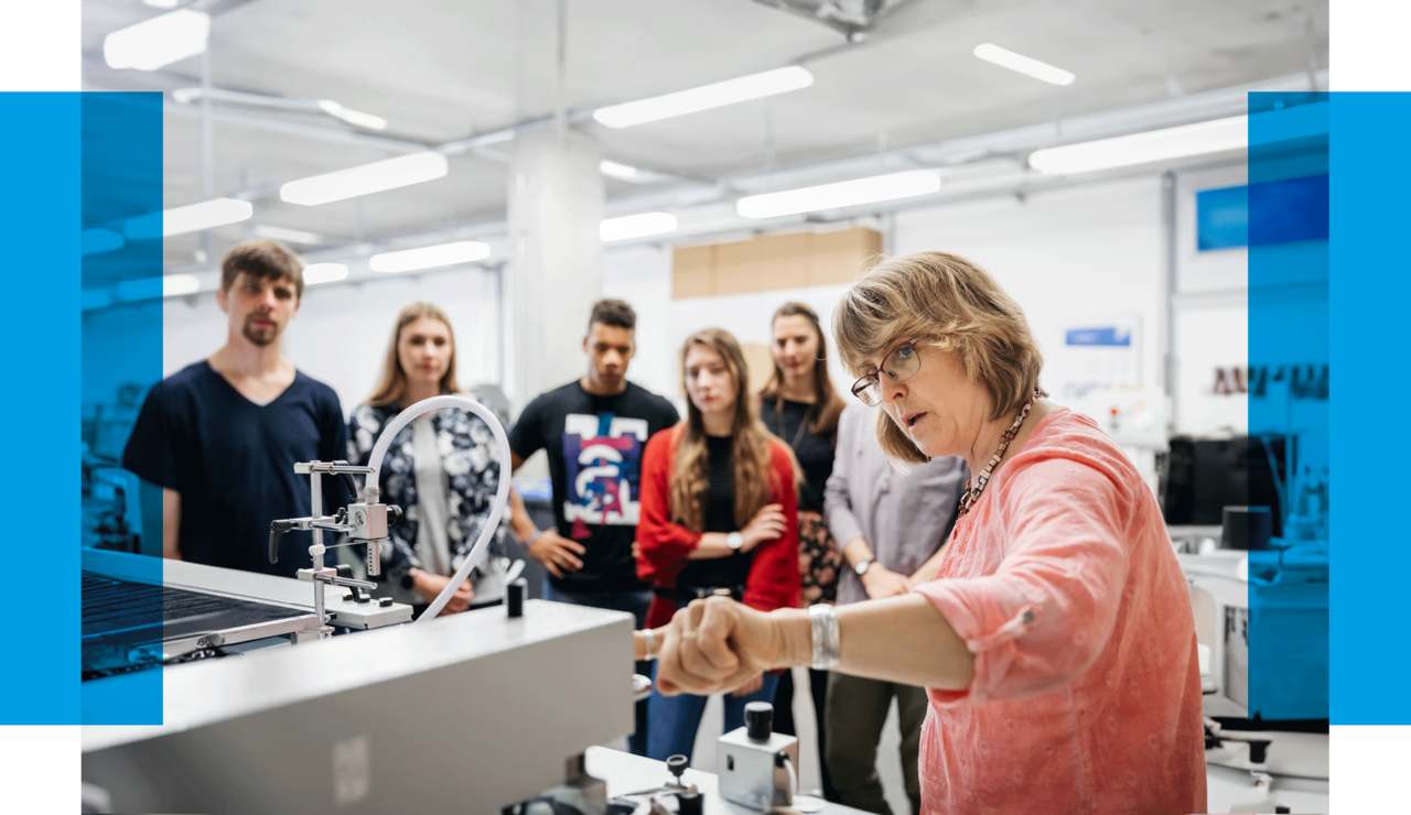 Eine Professorin erklärt Studierenden eine Druckmaschine. Dazu steht dort geschrieben: "Gestalten Sie die Zukunft mit!"