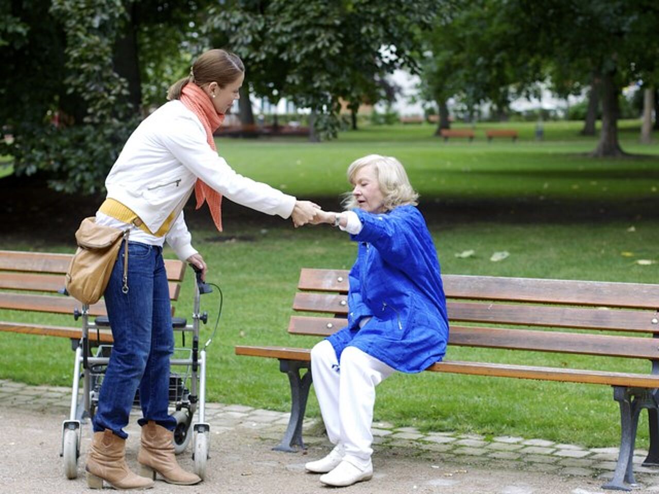 Schmuckbild zum Thema Pflege von Angehörigen. Eine jüngere Frau hilft einer älteren Frau beim Aufstehen von einer Bank in einem Park.