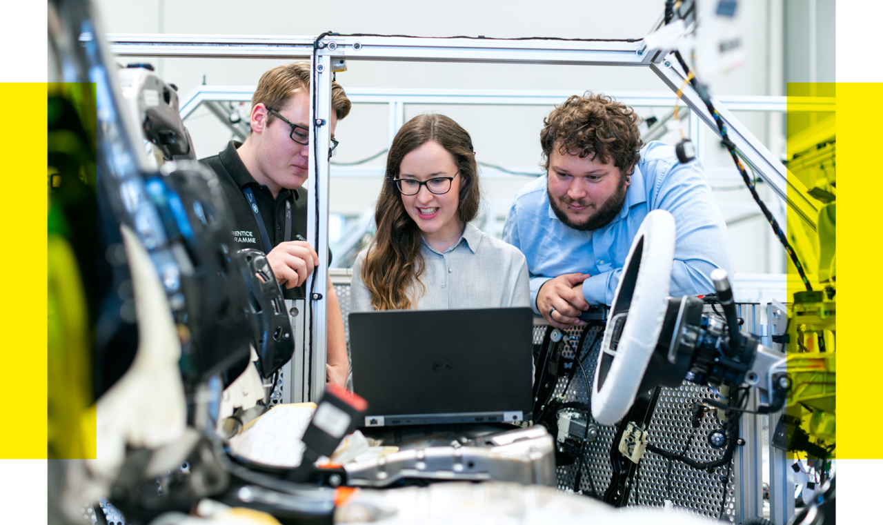 Drei junge Wissenschaftler:innen arbeiten gemeinsam an einem hochtechnologischen Experiment in einem Labor. Sie stehen um ein komplexes Gerät herum, dessen Teile und Kabel deutlich zu sehen sind. Eine Frau in der Mitte, mit einer Brille und einer grauen Bluse, blickt konzentriert auf einen Laptop-Bildschirm, während sie von ihren Kollegen flankiert wird. Ein Mann links im Bild trägt einen Pullover und eine Brille, und der andere Mann rechts trägt ein blaues Hemd. Alle scheinen in die Daten auf dem Bildschirm vertieft und diskutieren aktiv ihre Arbeit, umgeben von der fortgeschrittenen Laborausrüstung.
