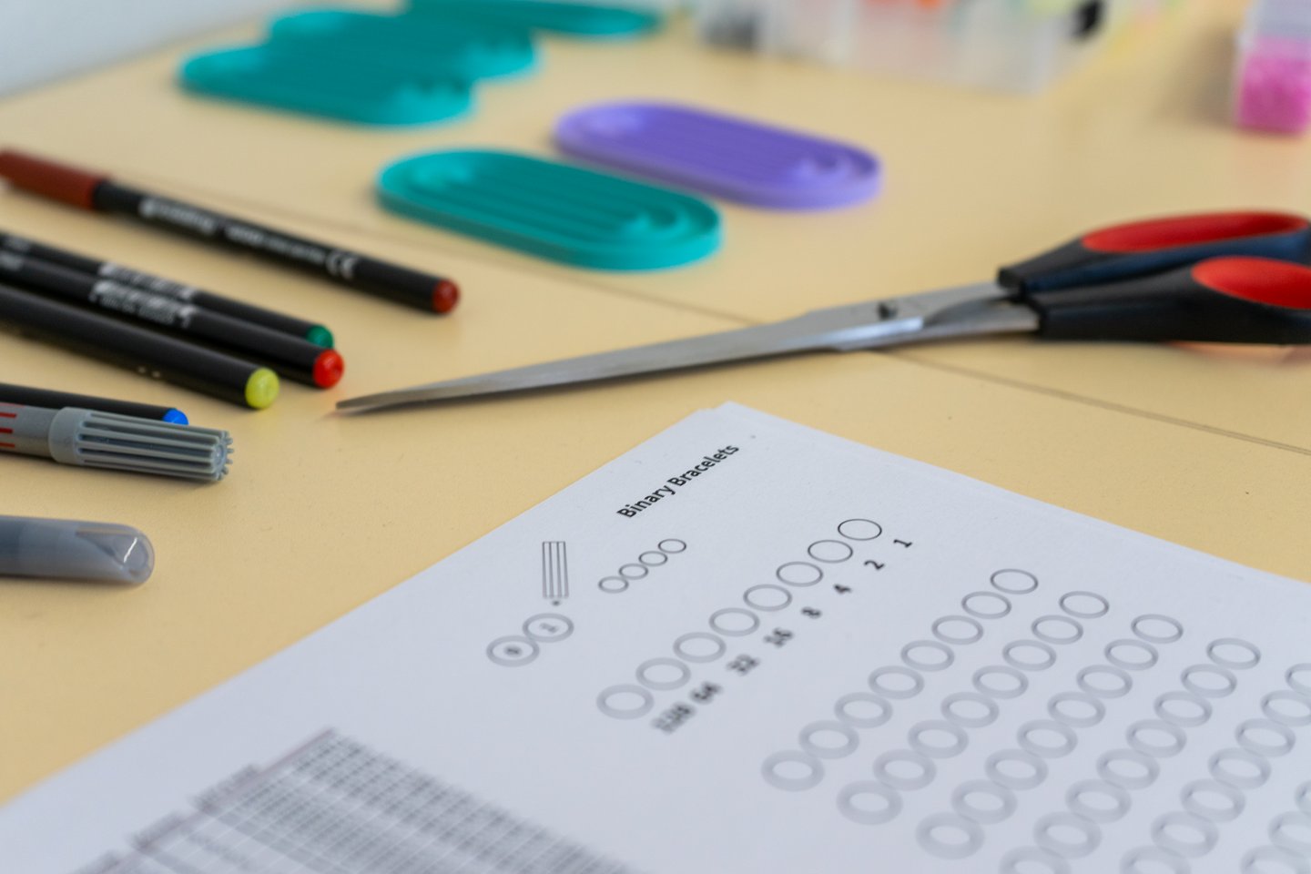 Workshop Binary Braceletts der Ferienhochschule für Girls im Winter 2026 Die Arbeitsutensilien bunte Filzstifte, Fädelbretter, Schere und Arbeitsblatt liegen auf einem Tisch.