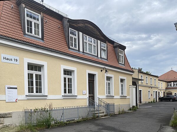 Klinikum St.Georg Haus 19 Das Bild zeigt einen Geweg dessen linke Seite mit Gebäuden versehen ist. Dabei sticht das nähste besonders ins Auge. Es handelt sich dabei um ein beiges Haus mit zwei Stockwerken. Die Hausfront der ersten Etage ist mit jeweils zwei Fenstern rechts und links ausgestattet und in dessen Mitte befindet sich die Tür. Diese werden von weißen Fenster- und Türfaschen umrandet. Links oben an dem äußersten Fenster befindet sich ein Schild mit der schwarzen Aufschrift "Haus 19". Darunter ist ein größeres Schild im Hochformat mit einer Beschriftung angebracht. Die Fensterbänke, sowie das Dach strukturieren mit weißen Gesims optisch das Gebäude. Das Dach ist mit braunen Ziegeln ausgestattet. Aus diesen ragen auf jeder Seite zwei Fenster empor und in dessen Mitte befindet sich ein großes, dreigesteiltes. Über diesem ragt eine halb-ovale Wölbung. Das Haus ist mit einem niedrigen Metallzaun abgegrenzt, der sich bis zum Treppengeländer der vier Stufen vor der Tür erstreckt.