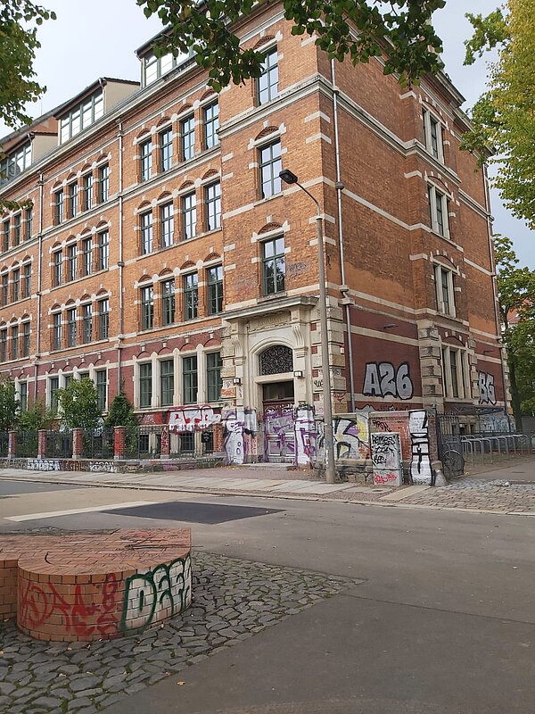 Es handelt sich um ein mehrstöckiges Gebäude aus roten Ziegelstein mit hellen Steinverzierungen. Der Eingangsbereich ist mit einem kunstvoll verzierten Bogen ausgestattet. Der untere Teil der Fassade ist stark mit Graffiti bedeckt.