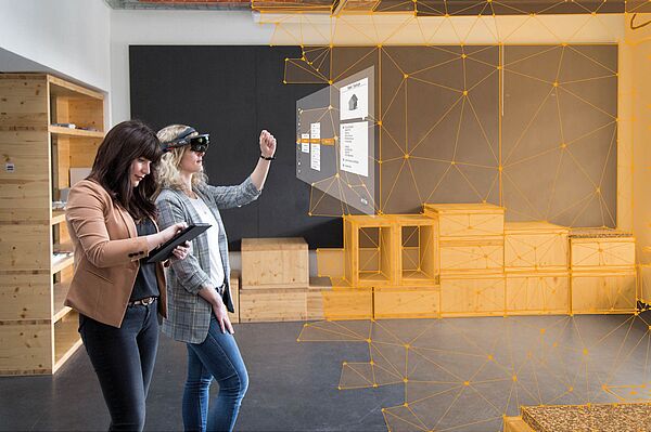 Sophie Seifert klickt auf ein Tablet, Lena Salm trägt eine HoloLens und klickt in die Luft  auf einen Bildschirm, der in der Luft schwebt. Im Hintergrund sieht man einen Raum, dessen Kanten digital nachgezeichnet wurden.