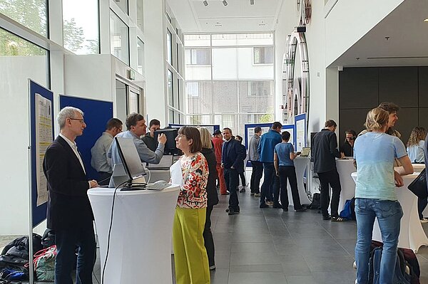 Menschen stehen in einem Foyer zusammen und unterhalten sich. Es sind Monitore und Pinnwände mit Postern zu sehen.