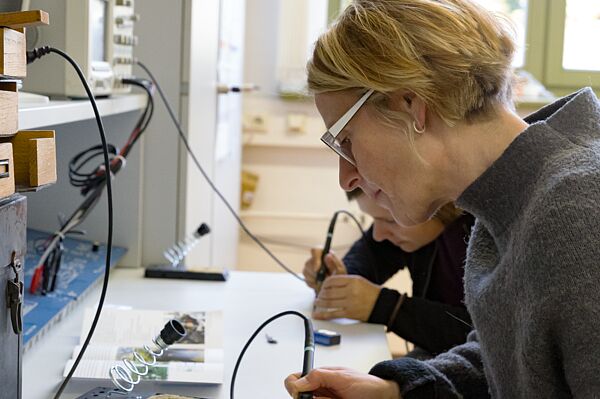 Hier entsteht gerade ein „Wasserwächter", den Birgit Uhlich im Unterricht von Laboringenieur Jan Dossin mit Hilfe des Lötkolben zusammenbaut. Eine Frau sitzt mit einem Lötkolben in der Hand vor einer Platine.