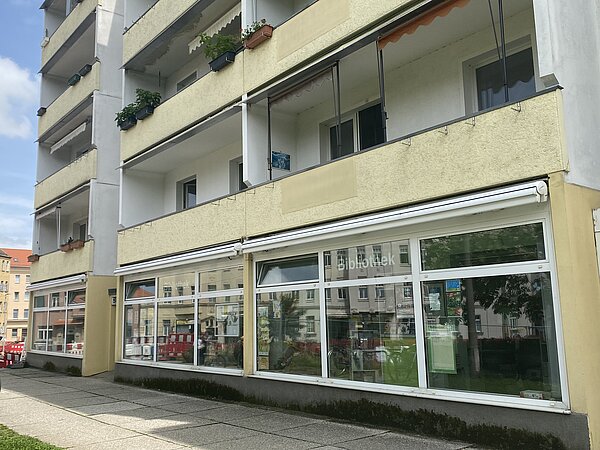 Der untere Teil eines weiß-gelben Plattenbaus, in dem sich die Stadtteilbibliothek Schönefeld befindet. Das Erdgeschoss hat viele große Fenster, in zwei Oberlichtern steht „Bibliothek“. 