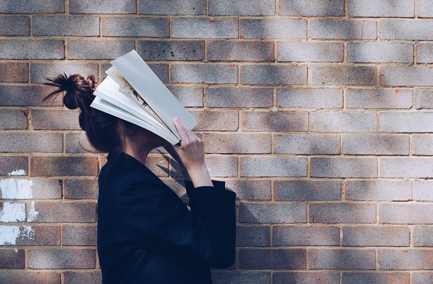 Symbolbild Lernstress Eine Frau schlägt ein Buch vor ihr Gesicht, während sie vor einer Hauswand steht,