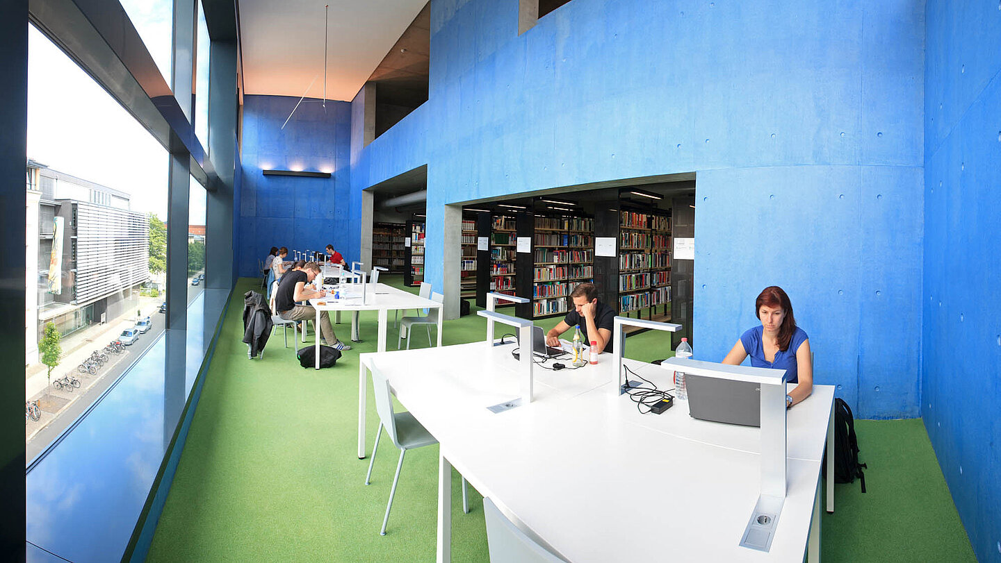 Bibliothek Innenansicht. Der Raum ist geräumig, es fällt viel Licht von einer Fensterwand ein. Die Wände sind in blau gestrichen. In der Mitte stehen mehrere weiße Tische an denen Menschen arbeiten. 