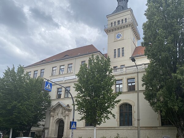 Friedrich-Schiller-Gymnasium Außenansicht eines sandfarbenen Gebäudes mit rotem Dach und braunen Fensterrahmen. Das Gebäude hat einen schmalen Turm mit einer Uhr und spitzem Dach auf der rechten Seite. Die Fassade ist an mehreren Stellen verziert. Vor dem Gebäude sind einige Bäume und ein Zebrastreifen Straßenschild.