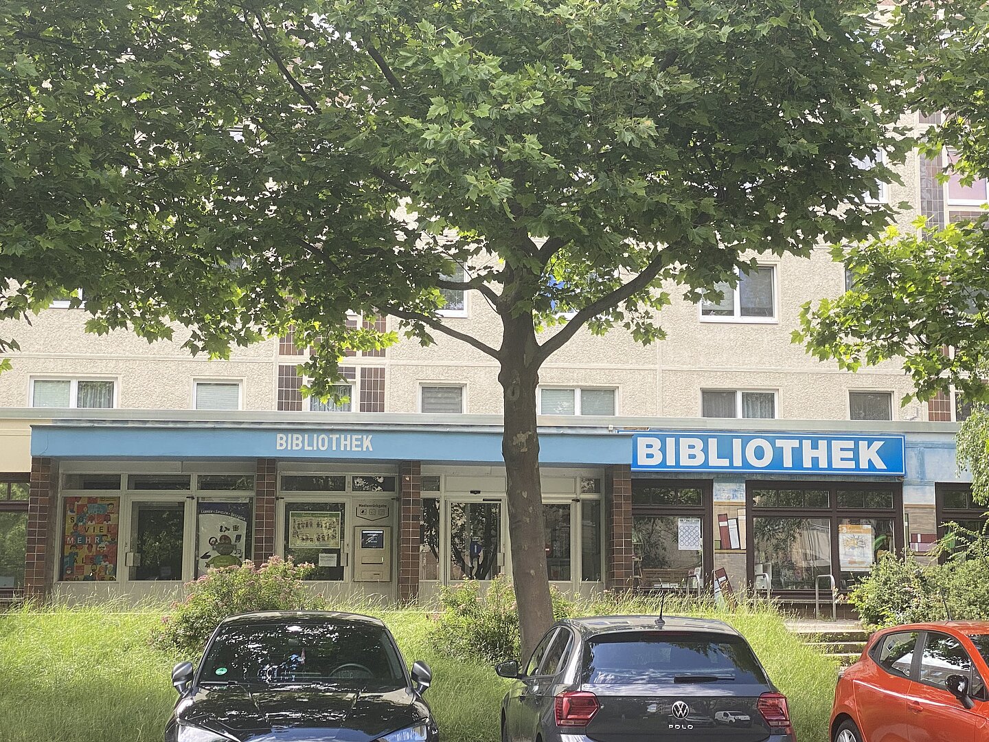 Leipziger Stadtteilbibliothek Paunsdorf Das Bild zeigt die Außenansicht einer Bibliothek. Die Fassade hat ein blaues Vordach, auf dem in weißen Buchstaben „BIBLIOTHEK“ steht. Es sind mehrere Fenster und Türen sichtbar. Vor dem Gebäude sind drei Autos geparkt, und ein Baum mit grünen Blättern verdeckt teilweise die Sicht. Über der Bibliothek sind Fenster von Wohnungen zu sehen. In den Fenstern der Bibliothek sind einige Plakate und Hinweise zu erkennen.