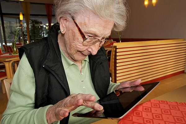 Ältere Person sitzt auf einem Stuhl und tippt auf ein Tablet. Im Hintergrund ist ein Essensraum.