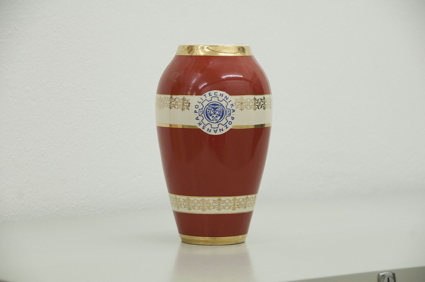 Vase von der TU Posen aus dem HTWK-Archiv (Foto: HTWK Leipzig) Zu sehen ist eine rote Vase aus Stein, die oben und unten einen vergoldeten Rand hat.