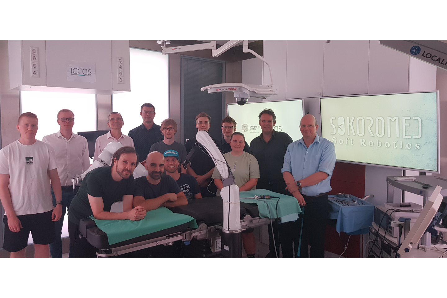 Research cooperation team in the ICCAS demo operating theatre, all looking into the camera, screens lit up in the background, operating table in the foreground