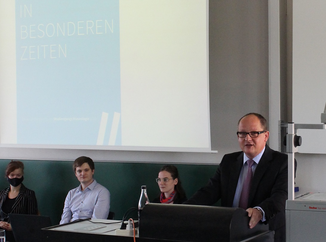 Ein Mann steht am Rednerpult, dahinter groß auf die Wand projiziert "In besonderen Zeiten"