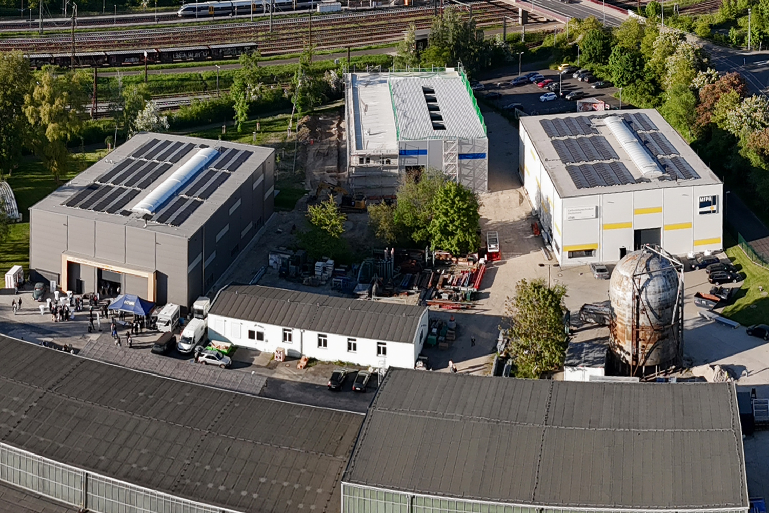 Drohnenaufnahme vom Campus mit mehrere großen Hallen in der Nähe von Wald und Schienen.
