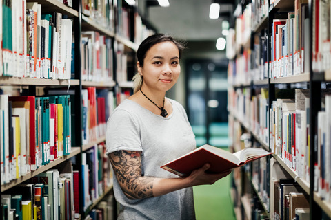 Eine Studentin steht mit Buch in einer Bibliothek.