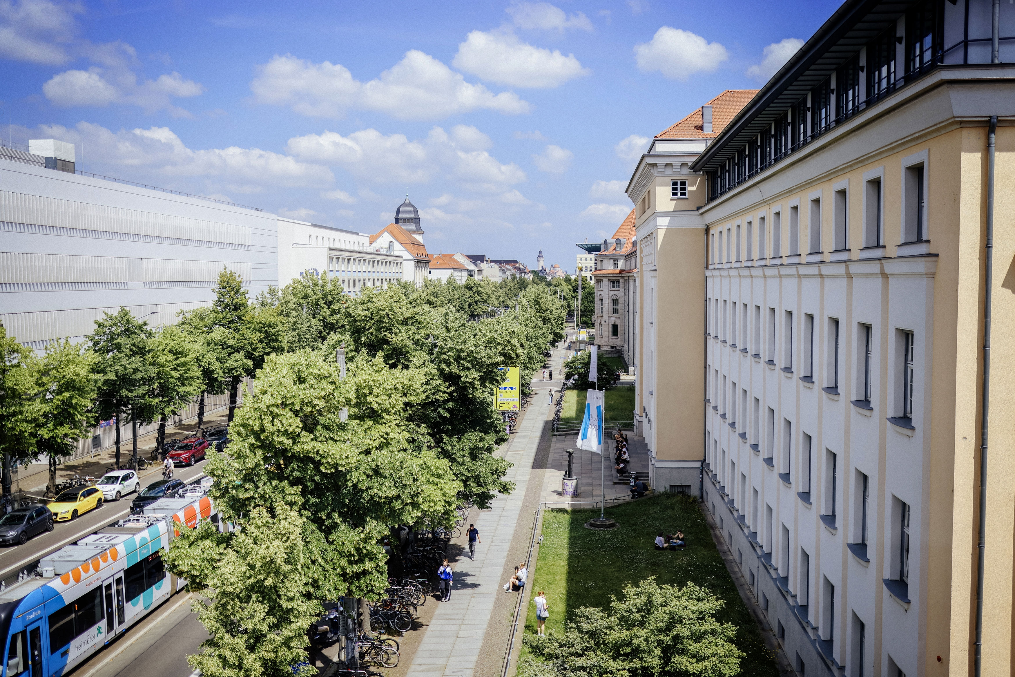 Blick auf eine belebte Straße mit Gehweg, Bäumen und historischen Gebäuden, Menschen sitzen und gehen, im Hintergrund Leipziger Stadtsilhouette.
