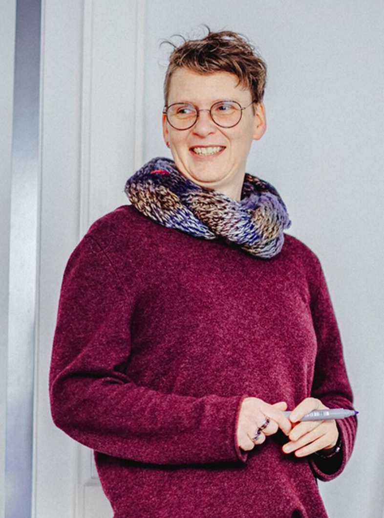 Person mit kurzem Haar trägt einen grob gestrickten Schal und einen dunkelroten Pullover und hält einen Stift in den Händen