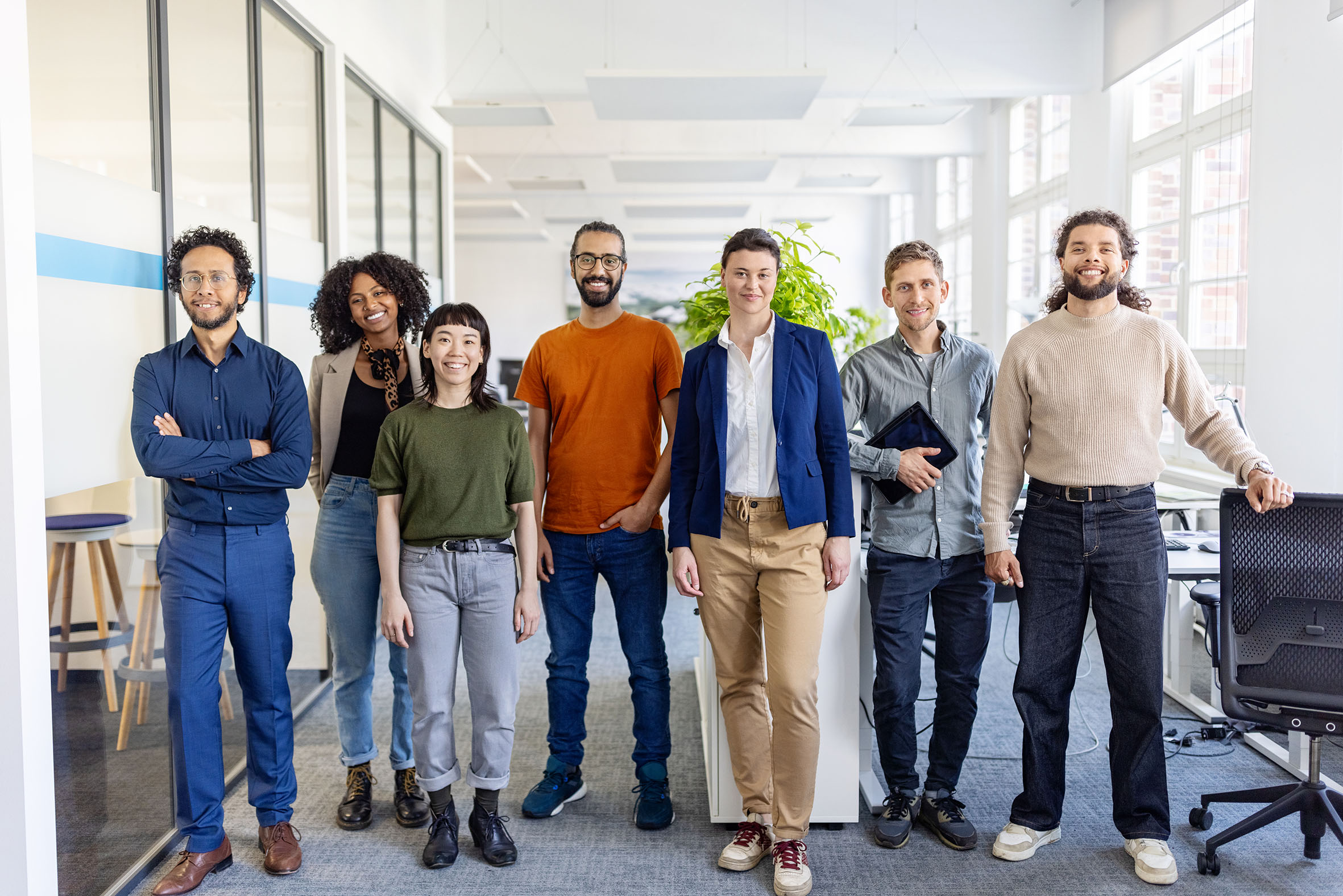 Eine multikulturelle Gruppe von jungen Erwachsenen steht in einem Büro und alle schauen frontal in die Kamera.
