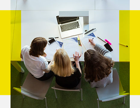 Drei junge Frauen aus der Vogelperspektive gesehen sitzen gemeinsam vor einem Laptop und schreiben Informationen mit.