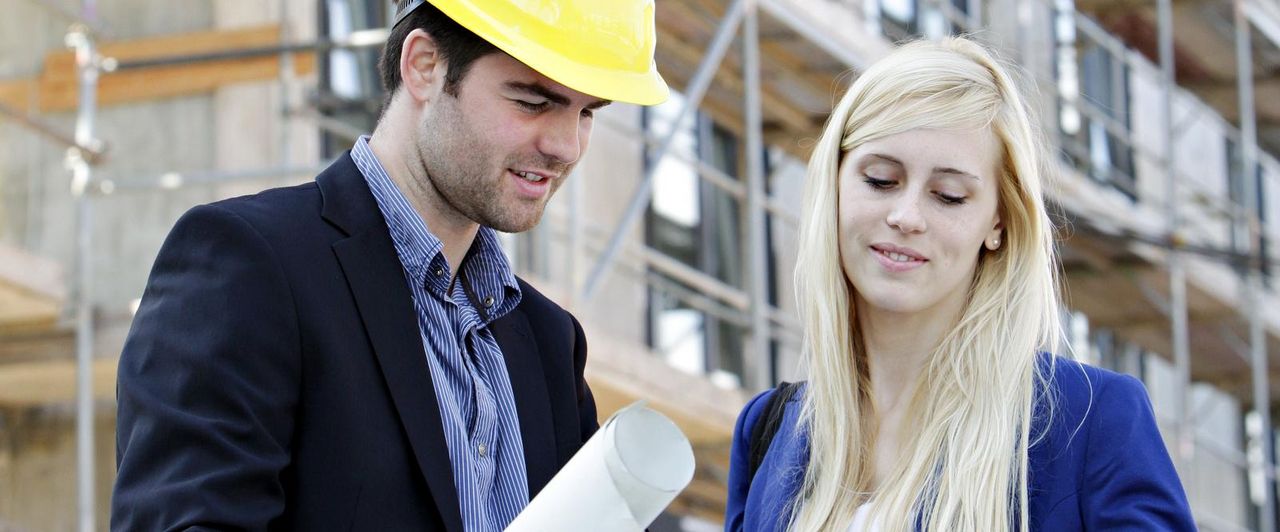 Halbtotale: Eine Frau und ein Mann stehen auf einer Baustelle und betrachten einen Bauplan.