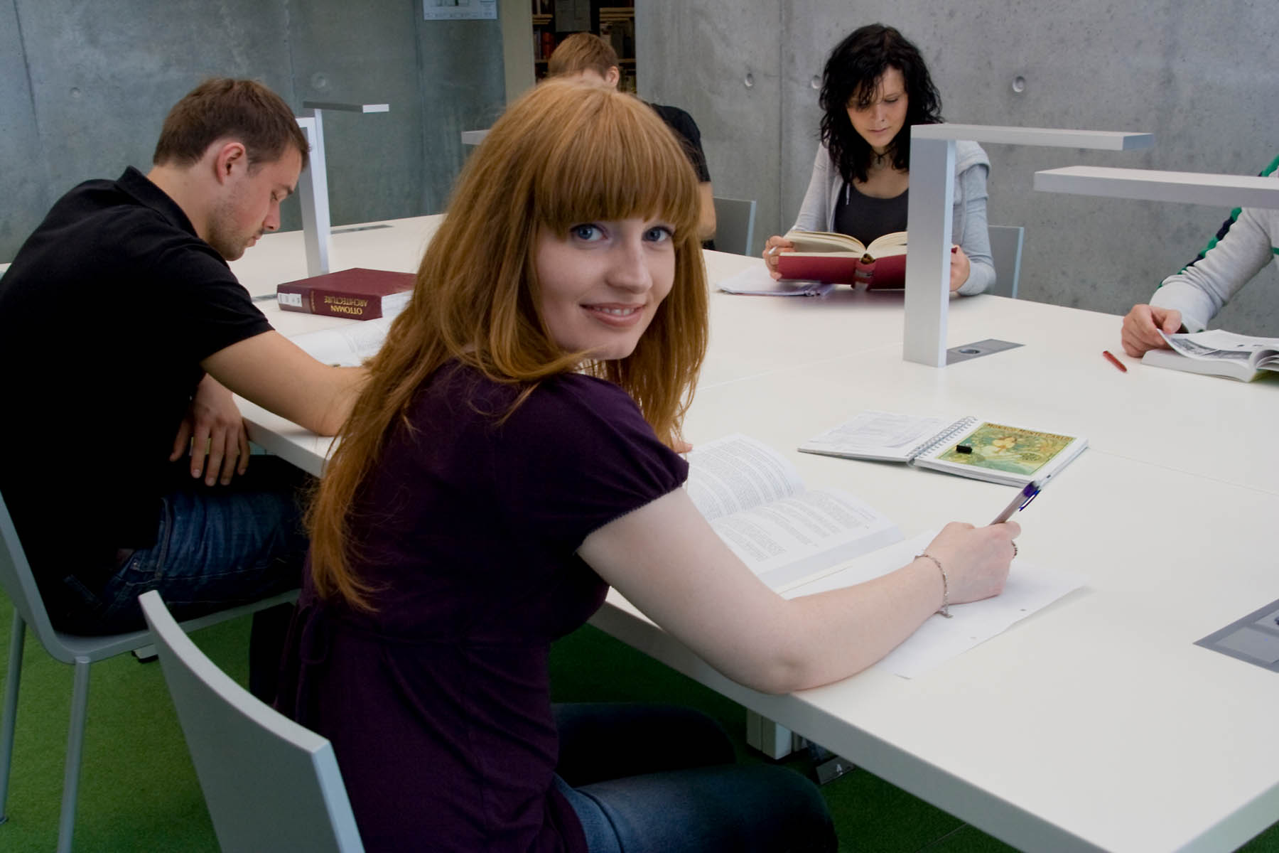 Studierende sitzen an einem Tisch in der Bibliothek in der HTWK. Die Studentin im Vordergrund ist zu Kamera hingewandt und lächelt.