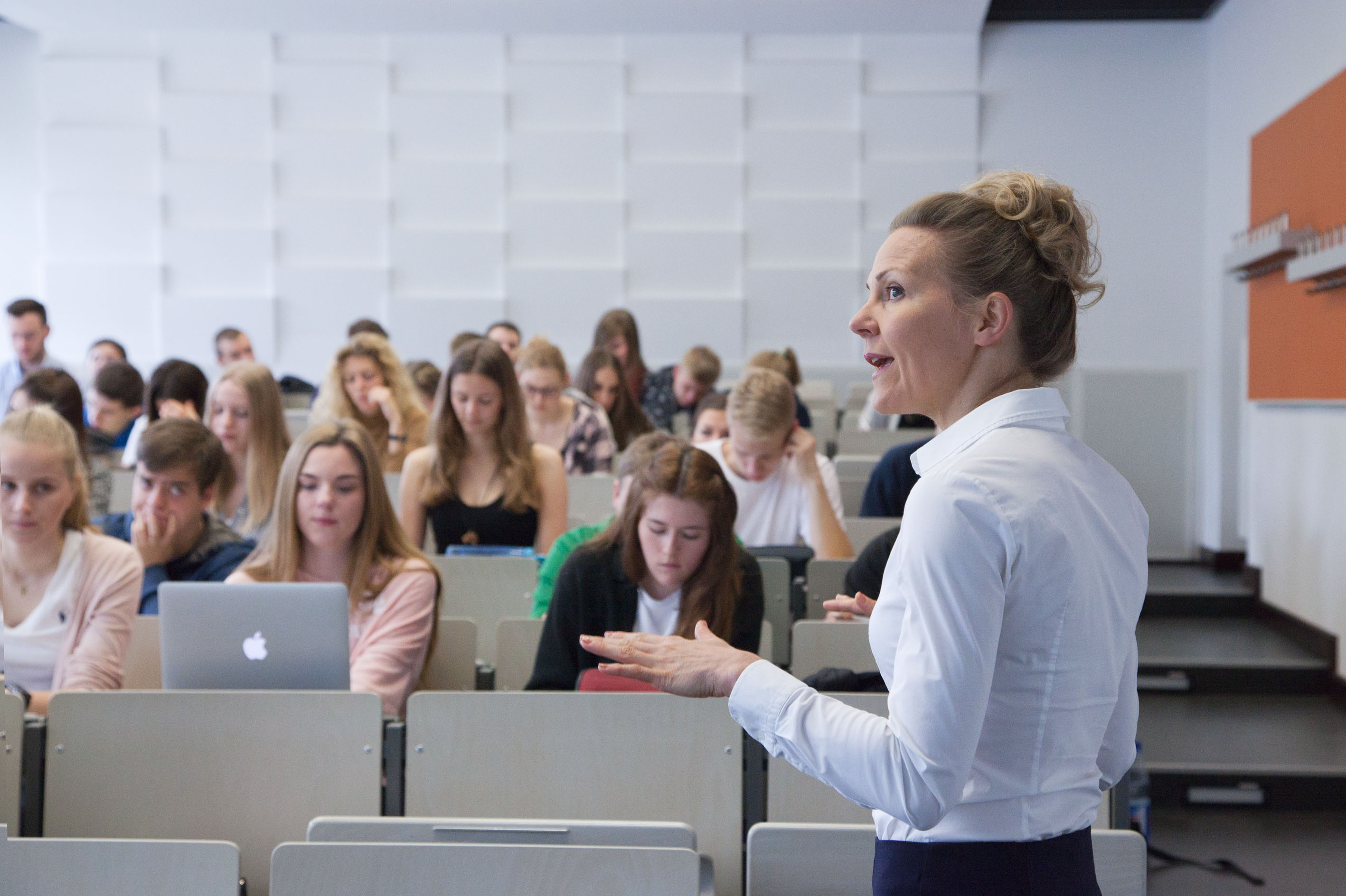 Blick auf eine Dozentin im Vordergrund, die den Studierenden im Hörsaal etwas erklärt. Oder: Ausschnitt eines Hörsaals mit Studierenden und einer Dozentin im Vordergrund.