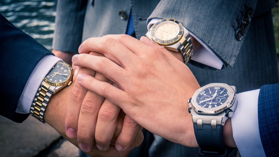 Abbildung dreier im Anzug gekleideter Personen, deren Hände übereinander liegen. Alle drei Personen tragen eine große Armbanduhr.