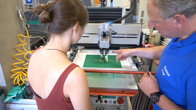 Schnittbild von einem Dozenten und einer Studentin, die zusammen an einer Druckmaschine arbeiten. 