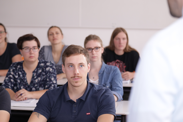 Frontalaufnahme von Studierenden, die an Tischen sitzen. Fotografiert aus der Dozentensicht.