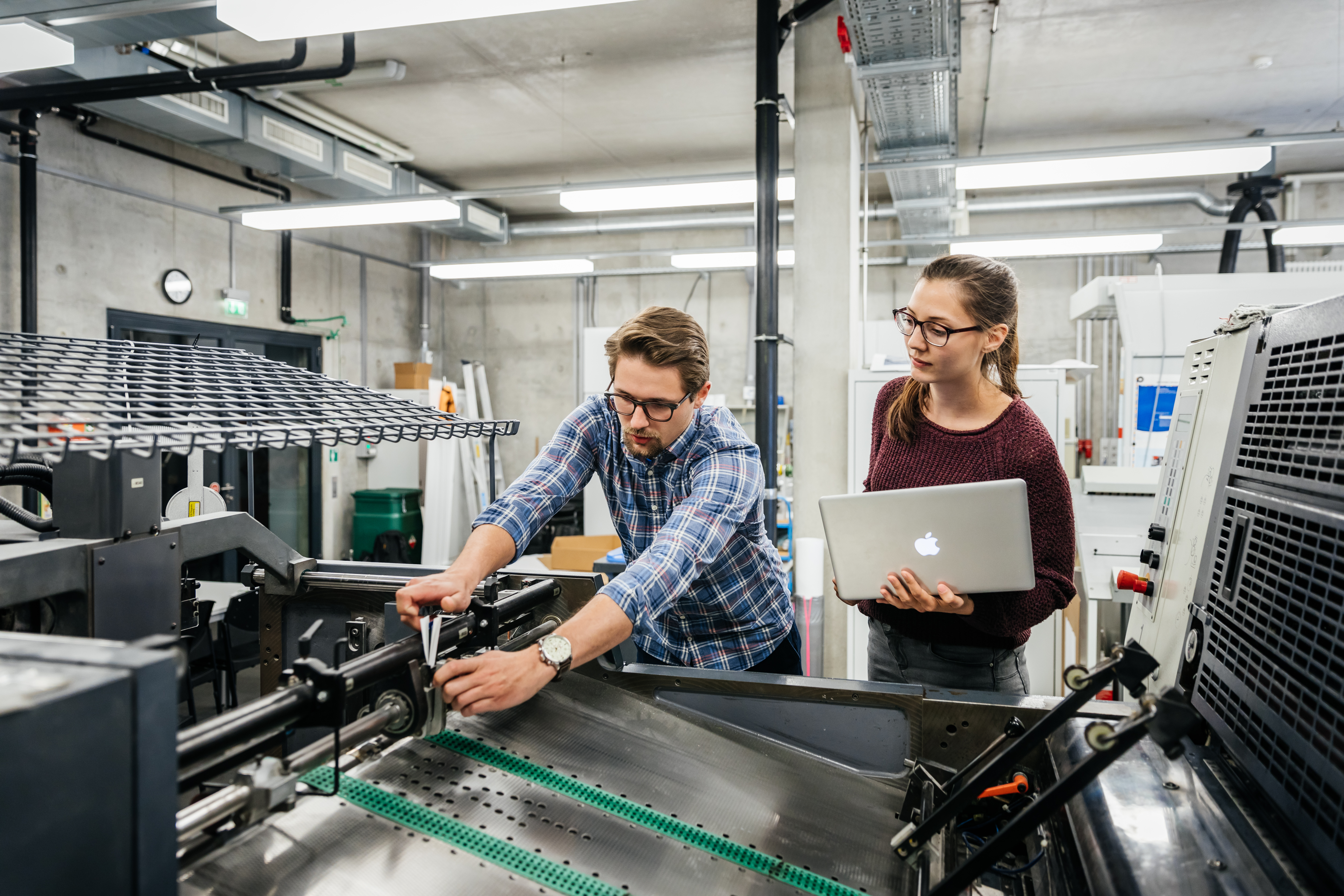 Ein junger Mann beught sich über eine Maschine und eine junge Frau mit Laptop schaut ihm über die Schulter