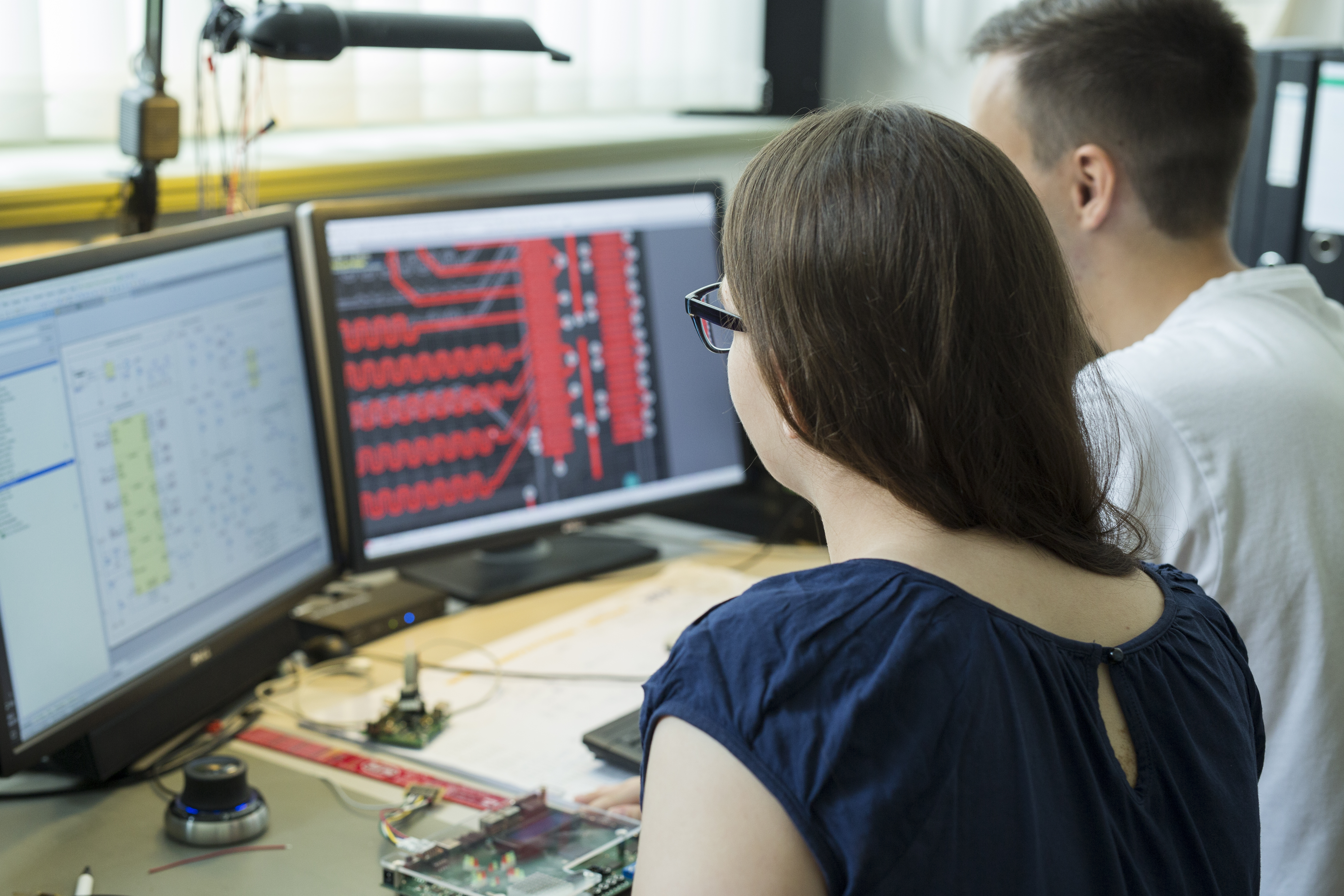 Auf dem linken Monitor sieht man einen Schaltkreisentwurf und auf dem rechten Monitor ein Leiterplattenlayout. Auf dem Schreibtisch steht eine Platine mit elektronischen Bauteilen.