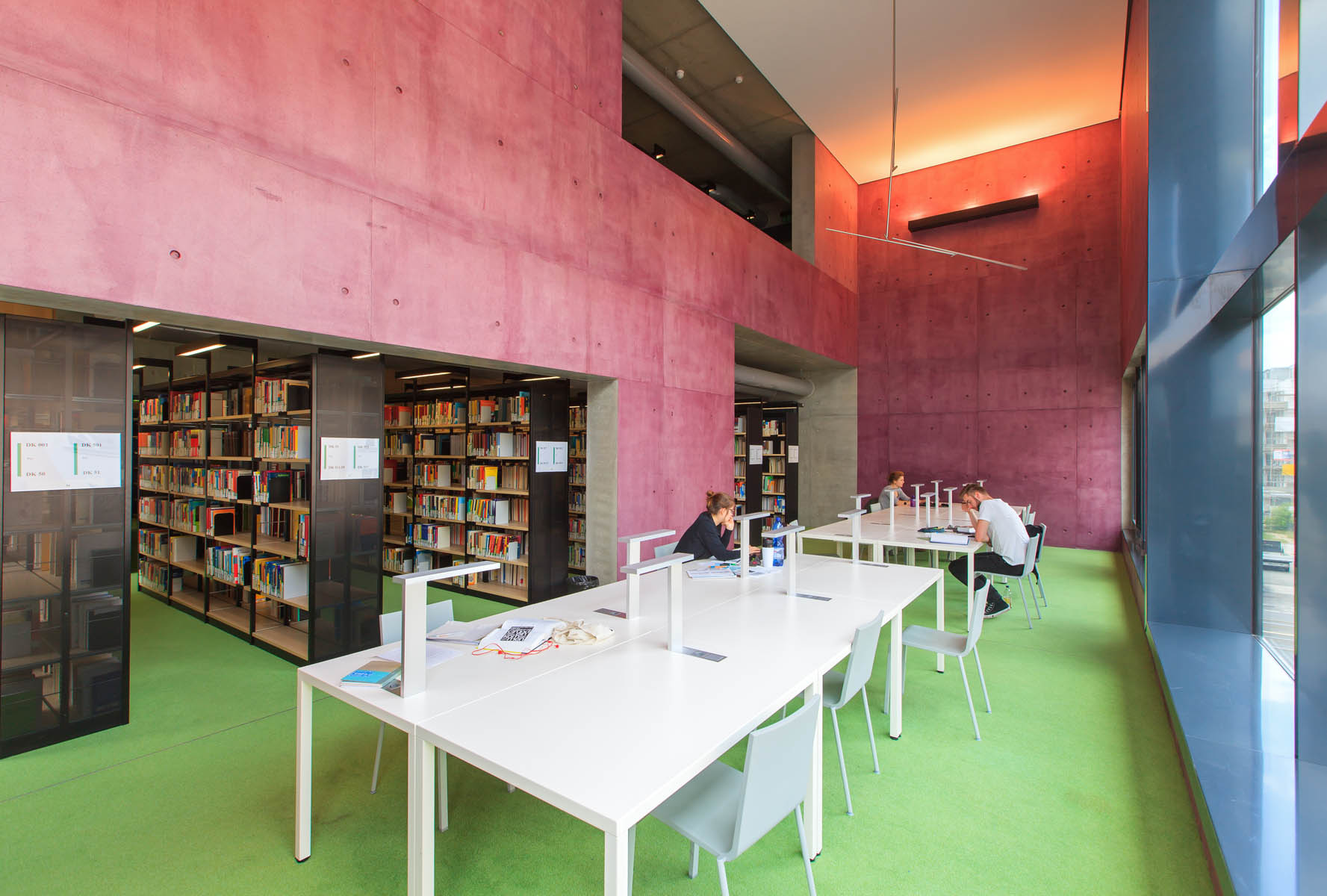 Bibliothek Innenansicht. Der Raum ist geräumig, es fällt viel Licht von einer Fensterwand ein. Die Wände sind in roter Farbe gestrichen. In der Mitte stehen mehrere weiße Tische an denen Menschen arbeiten.