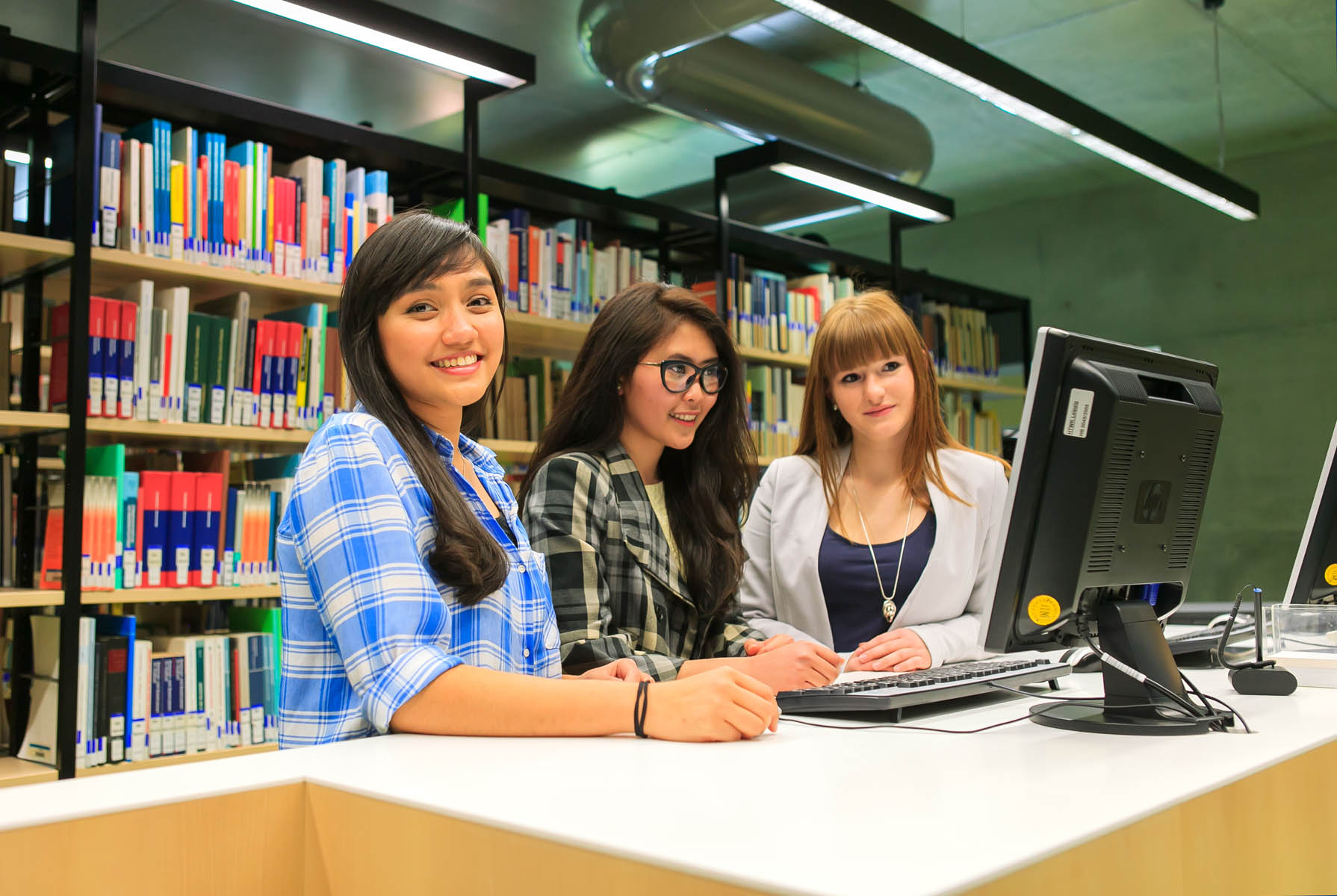 Drei Studentinnen sitzen an einem Schreibtisch vor einem Computer. Im Hintergrund stehen Bücherregale. Die Studentin links schaut in die Kamera.
