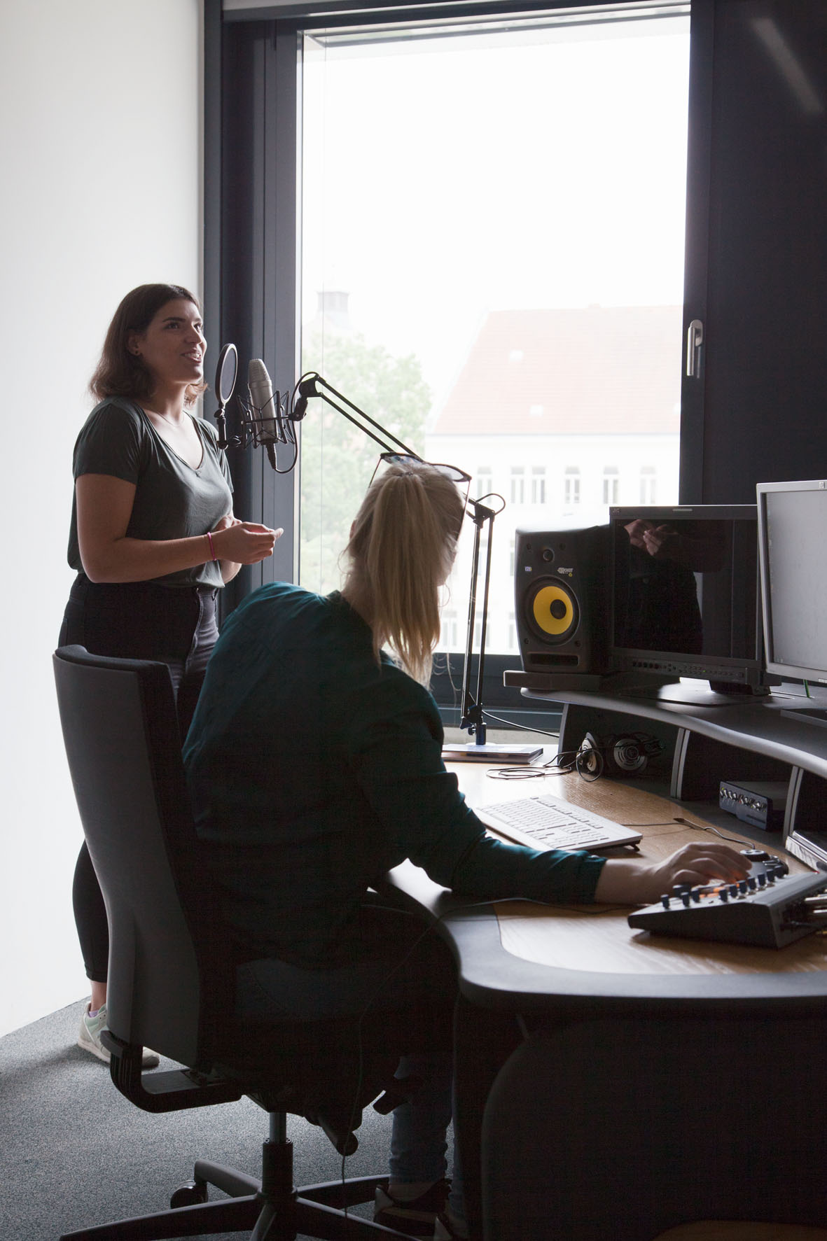 Szene im Aufnahmestudio der HTWK: Eine Studentin spricht in ein Mikrofon während eine andere Studentin, die am Schreibtisch sitzt, die Regler eines Mischpults betätigt.