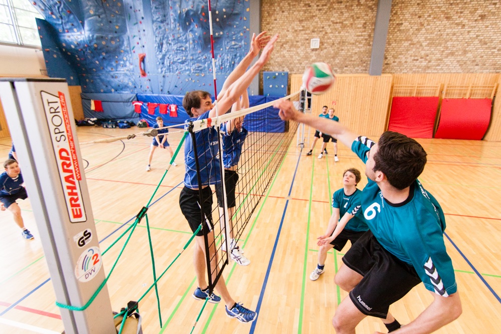 Mehrere Studenten spielen Volleyball. Jeweils zwei Studenten befinden sich direkt am Netz und versuchen den Ball zu blocken.