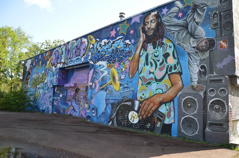 Foto einer Häuserwand, die mit Grafiti besprüht ist. Die Grafiti zeigen unter anderem einen DJ, der an den Turntables steht, einen Breakdancer, mehrere große Musikboxen und Trompeten.