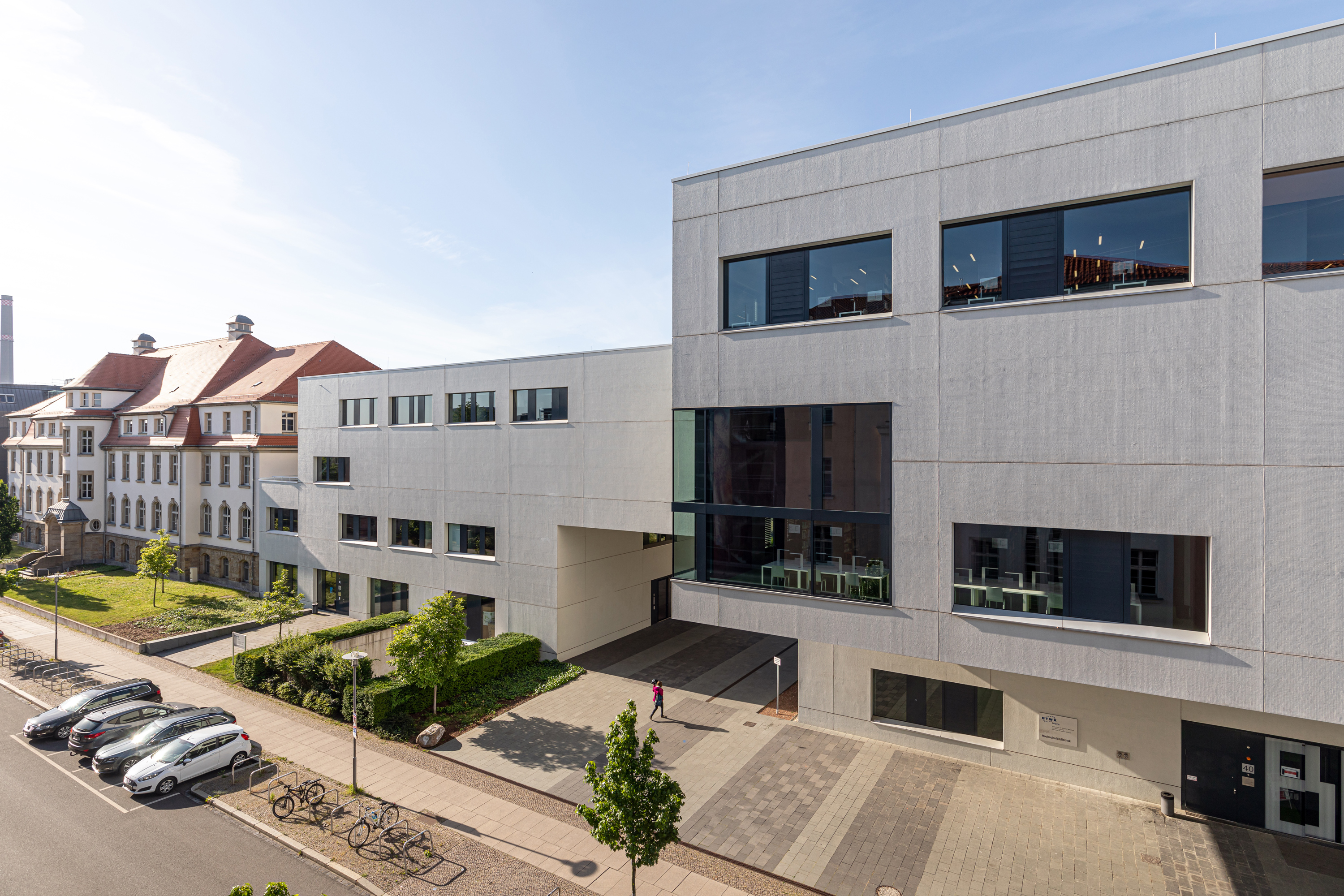 Hochschulbibliothek und Medienzentrum Im Vordergrund zwei weiße, moderne kubische Gebäude an einer Straße. Im Hintergrund ein älteres Hochschulgebäude.